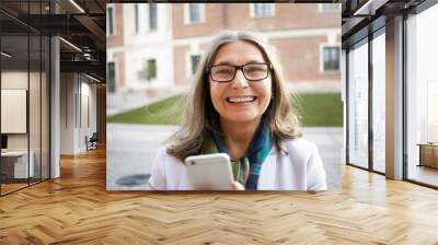 Horizontal shot of beautiful middle aged female pensioner with gray hair posing on city street, holding modern electronic device, looking at camera with happy broad smile. Age and technology Wall mural