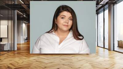 Horizontal studio portrait of adorable beautiful young overweight European woman with loose black hair and chubby cheeks wearing round earrings and xxl white shirt posing at blank wall, smiling Wall mural