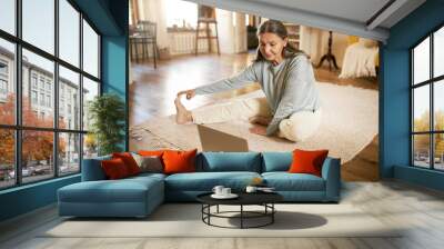 Indoor image of flexible active mature gray haired female sitting on floor in front of open laptop, stretching legs during online yoga lesson, watching live video with professional instructor Wall mural