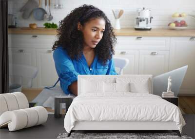 Indoor shot of attractive serious young dark skinned with wavy hair doing paperwork in kitchen, typing numbers in online form, holding paper bill, paying for gas, electricity and rent using laptop Wall mural