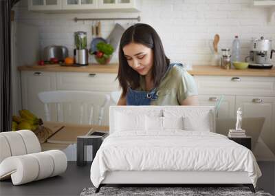 Indoor shot of attractive young female doing calculations, managing home budget, sitting at kitchen table and smiling. Pretty girl paying domestic bills online, using laptop computer and calculator Wall mural