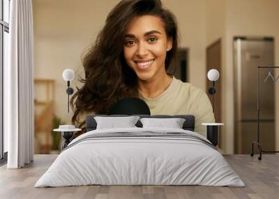 Indoor shot of beautiful happy young 20 year old Arabic woman relaxing at home wearing comfy clothing, looking at camera with broad smile, showing white straight teeth. Cute girl having rest indoors Wall mural