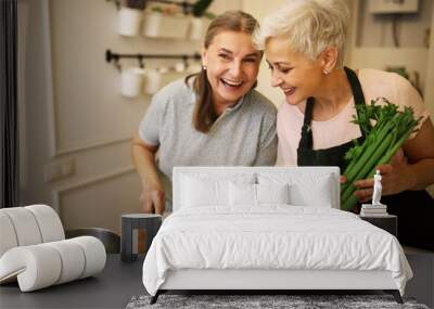 Indoor shot of charming retired sisters cooking lunch together at home waiting for guests. Cheerful mature woman in apron holding celery, cooking vegetable soup with her best friend, laughing Wall mural