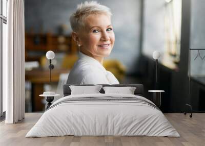 Indoor shot of fashionable successful mature blonde woman in formal white blouse turning head to camera with welcoming friendly smile, giving tour around her office. People, age, career and employment Wall mural