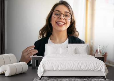 Indoor shot of friendly confident millennial female vlogger in stylish round eyeglasses speaking at camera, making live video for vlog on her social media account. People and lifestyle concept Wall mural