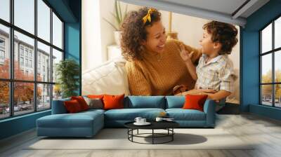 Indoor shot of happy young Hispanic woman with brown wavy hair relaxing at home embracing her adorable toddler son. Cheerful mother bonding with infant son, sitting on sofa in living room, laughing Wall mural