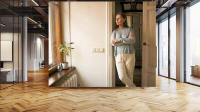 Indoor shot of serious sad middle aged female in casual clothes standing at doorway keeping arms crossed on her chest, looking through window, being upset while staying at home during quarantine Wall mural