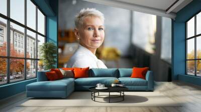 Isolated image of beautiful stylish middle aged female entrepreneur with neat make up going to business meeting, standing against office interior background, looking at camera with confident smile Wall mural