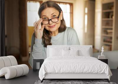 Mature age, people and distant work concept. Attractive confident middle aged woman in glasses working from home, sitting in front of open laptop, doing paperwork, looking at camera with happy smile Wall mural