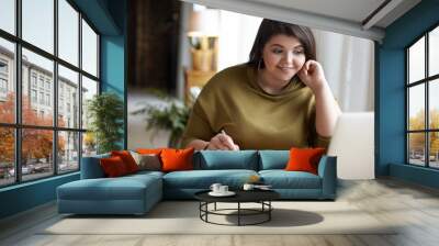 Modern electronic gadgets, job and occupation concept. Cute elegant young brunette female with excess weight using laptop computer for remote work, looking at screen, holding pen, writing down Wall mural