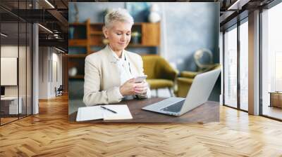 Modern lifestyle, communication and electronic gadgets concept. Busy attractive elderly female entrepreneur with pixie blonde hair working in office with laptop and notes on desk, using smart phone Wall mural