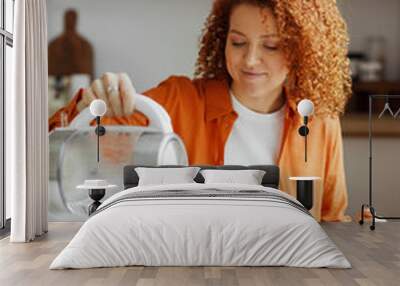 Morning breakfast. Happy smiling woman with red curly hair pouring hot water from transparent kettle in cup with tea, sitting at kitchen table in front of bowl with fruit salad or muesli Wall mural