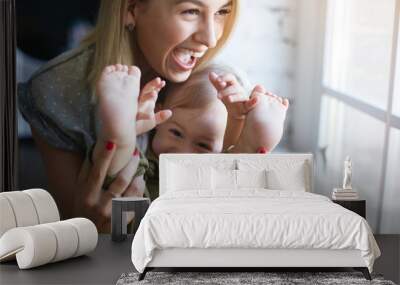 People, family, happy childhood, joy and love concept. Cheerful attractive young woman holding feet of her cute baby son, opening mouth widely in excitement, having fun indoors. posing by window Wall mural