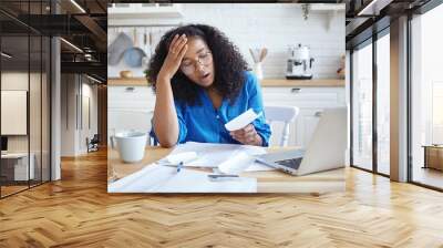 People, modern technology, finance and housework concept. Picture of tired young dark skinned woman in eyeglasses doing paperwork at home, having frustrated look, shocked with amount of debt Wall mural