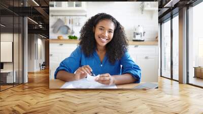 People, paperwork and modern technology concept. Picture of joyful young African woman holding pen and bill while calculating family budget at home, sitting in kitchen with papers and laptop on table Wall mural