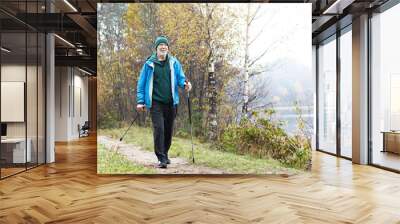 Picture of active fit Caucasian elderly man wearing sports clothes and warm hat having walk using nordic poles outdoors. Retired senior male nordic pole walker enjoying autumn morning outside Wall mural