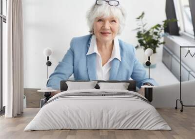 Picture of attractive confident elderly mature female financial adviser with short gray hair looking at camera with smile, studying piece of paper in her hands while working at her office desk Wall mural