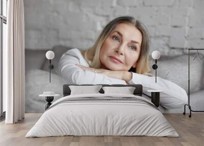 Picture of attractive middle aged Caucasian woman with long straight hair resting on grey comfortable sofa, having sad unhappy expression, feeling bored or lonely. People, lifestyle and age concept Wall mural