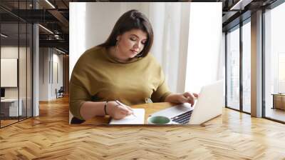 Picture of attractive young European female journalist wearng stylish earrings and knitted sweater making notes in diary while working on research for her new article, looking for information online Wall mural