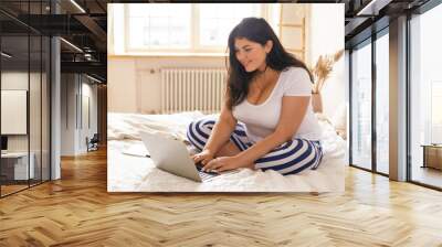 Plump overweight young woman in striped trousers asitting comfortably on bed with legs crossed, using laptop to chat and make new friends online via social network or dating website, smiling Wall mural