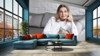 Portrait of attractive positive middle aged blonde Caucasian woman in her sixties spending leisure time indoors, resting on couch in living room, looking at camera with pleased joyful smile Wall mural