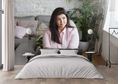 Portrait of attractive young overweight dark haired woman daydreaming indoors, planning her day, sitting on gray sofa surrounded with many pillows, dressed in stylish pink shirt and leggings Wall mural