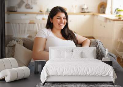 Portrait of cheerful charismatic young overweight female with big breast and long black hair posing in stylish kitchen interior, having coffee on comfortable couch and laughing at funny joke Wall mural