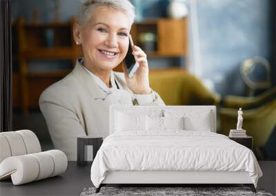 Portrait of confident short haired 60 year old female entrepreneur in beige formal suit posing in modern office interior using mobile making phone call, having business talk to partner, smiling Wall mural