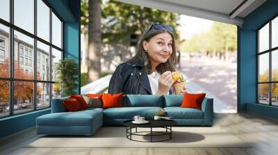 Portrait of fashionable mature European woman eating tasty ice cream cone, sitting outdoors on bench in park, having pleased joyful facial expression, her look expressing delight, saying Yummy Wall mural