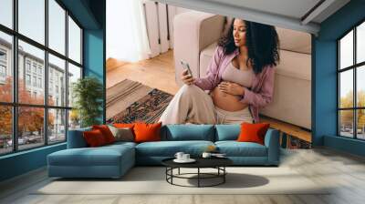 Portrait of gorgeous pregnant female with dark skin and curls sitting on carpet next to couch browsing news feed in her social media app, reading article, watching podcast for future moms Wall mural