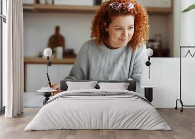 Portrait of happy female student watching webinar or tutorial studying online on laptop sitting at kitchen table, noting down after teacher or lecturer, looking at screen with interest Wall mural