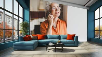 Portrait of happy smiling elderly female with gray hair in stylish shirt and glasses talking on phone leaning against wall, enjoying nice conversation with best friend living abroad Wall mural