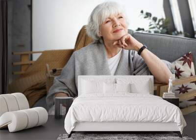 Portrait of lovely middle aged grey haired European woman with dreamy smile and eyes full of wisdom relaxing at home alone, sitting on comfortable couch, reminiscing about days of her youth Wall mural