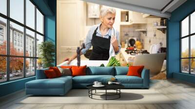 Portrait of middle aged female posing in kitchen wearing apron using laptop while cooking dinner, watching video recipe. Mature woman washing vegetables to make salad, following online instructions Wall mural