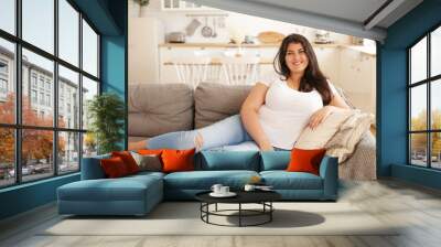 Portrait of positive young overweight brunette female sitting barefooted on gray sofa with legs outstretched wearing ripped jeans and white t-shirt looking at camera with happy cheerful smile Wall mural