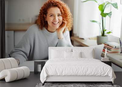 Portrait of smiling redhead curly female student studying at home, doing home work in front of laptop, preparing for exams, sitting at kitchen table with pen in hands, looking at camera Wall mural