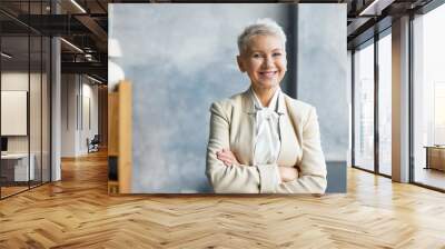 Portrait of successful attractive senior businesswoman with stylish short hairdo and confident smile posing in her modern office, keeping arms crossed on chest, wearing elegant beige formal suit Wall mural