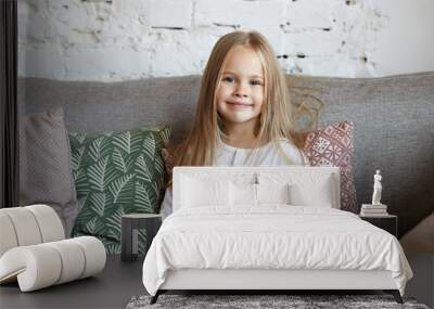 Positive beautiful little girl of European appearance sitting on comfortable sofa in living room, surrounded with decorative pillows, looking at camera and smiling happily. Childhood concept Wall mural