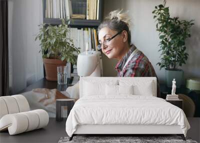 Positive casually dressed mature female with hair knot smiling pleasantly, enjoying process of creation of garment, sitting at desk, using sewing machine, surrounded with decorative plants and books Wall mural