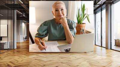 Pretty elderly bossy stylish female HR manager working at her office in front of laptop, signing papers or reading CV of new candidate on vacant position in their company, looking at camera smiling Wall mural