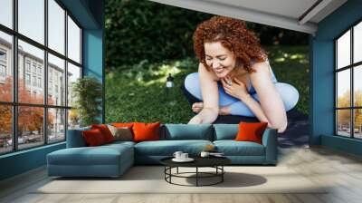 Pretty ginger head girl in sportswear sitting in park in front of laptop on mat next to bottle of water, putting hand to chest showing gesture of gratitude to her fitness tutor for online yoga Wall mural