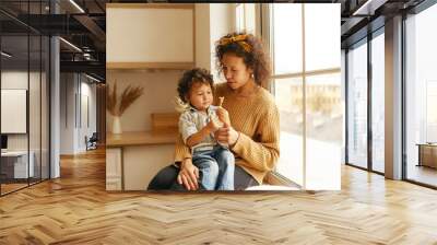 Pretty woman with curly hair sitting on windowsill with adorable baby on her lap, giving him toy or candy, little child looking with interest and curiosity. Motherhood, childcare and togetherness Wall mural