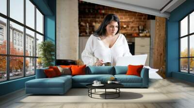 Serious plus size young businesswoman in white blouse working distantly from home office checking email, doing paperwork, sitting at kitchen table with laptop pc, documents and cup of coffee Wall mural