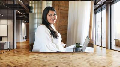 Side view of confident skillful young overweight female copywriter or translator sitting at table in front of open laptop, using wireless internet connection for distant work, smiling, drinking coffee Wall mural