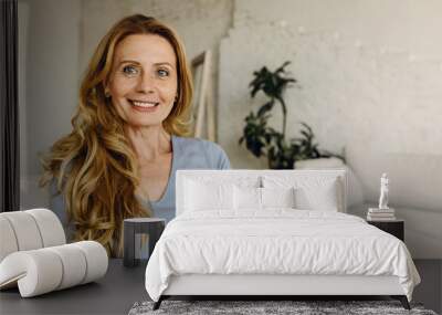 Smiling mature woman sitting on sofa against white home interior background, looking happy, smiling widely, looking at camera, having rest after house work, dressed casually in blue clothes Wall mural