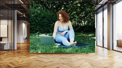 Smiling redhead barefoot female yoga instructor in blue leggings and top sitting on mat, greeting her followers in the begging of online workout tutorial, setting up good internet connection on laptop Wall mural