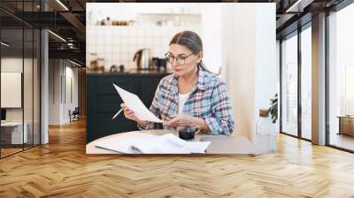 Stylish serious elderly Caucasian woman in spectacles and plaid shirt studying paper in her hand while working through papers in kitchen. Attractive mature female calculating gas and electricity bills Wall mural