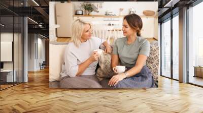 Sweet cozy portrait of beautiful mature female with blonde dyed hair holding cup, having coffee with her charming young daughter talking, sharing secrets or gossips, sitting on couch in living room Wall mural