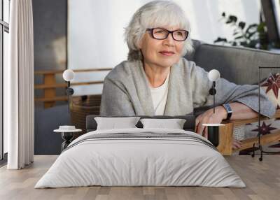 Thoughtful middle aged mature grey haired female wearing eyeglasses, wide scarf and wrist watch spending leisure time at home, sitting on comfortable sofa in living room, having pensive look Wall mural