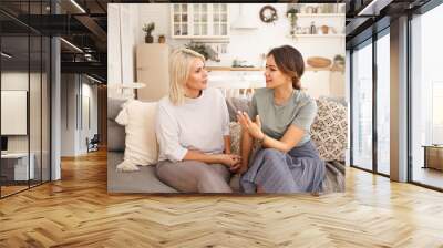 Two Caucasian women having nice lively conversation in living room. Cute young woman gesturing emotionally, proving something while talking to her middle aged mother on sofa in cozy interior Wall mural